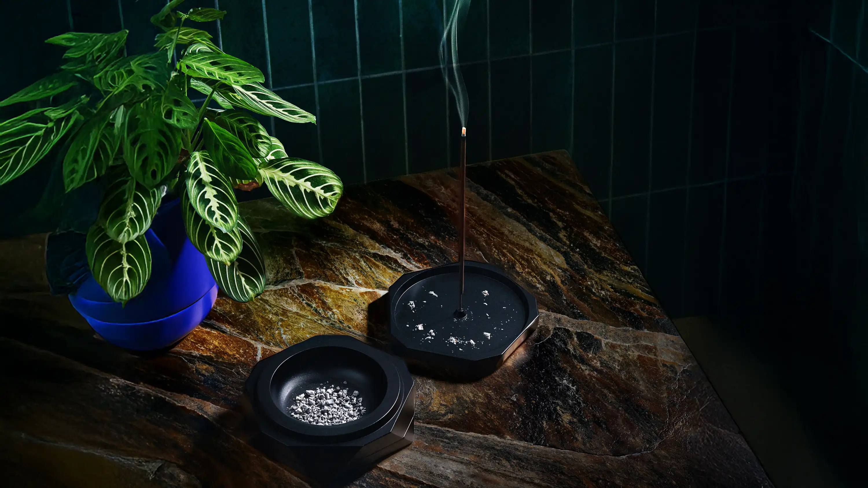 Black Vesta magnetic vessel used as ashtray and incense holder on a stone surface with rising smoke and a green plant in a blue pot against dark tiled background