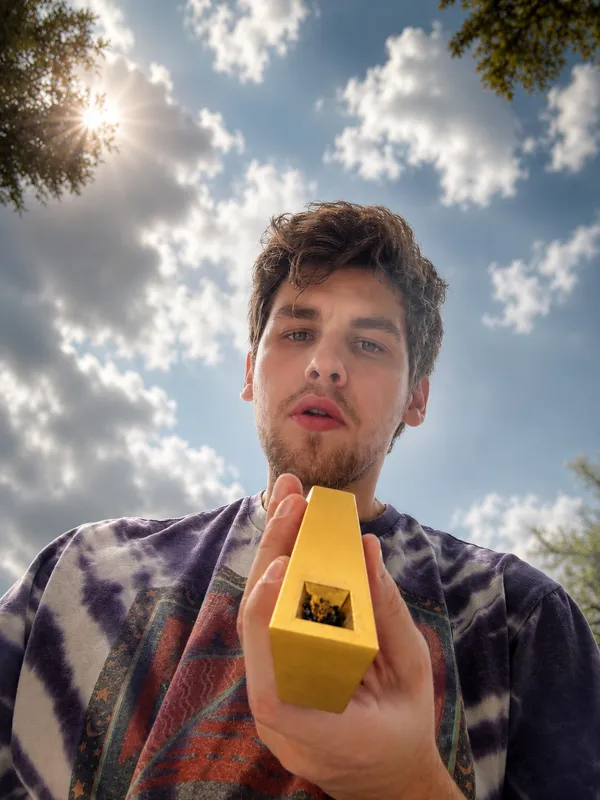 A young man holding a gold Tempest Radiance pipe that is still smoking after he took a hit outdoors under cloudy sky