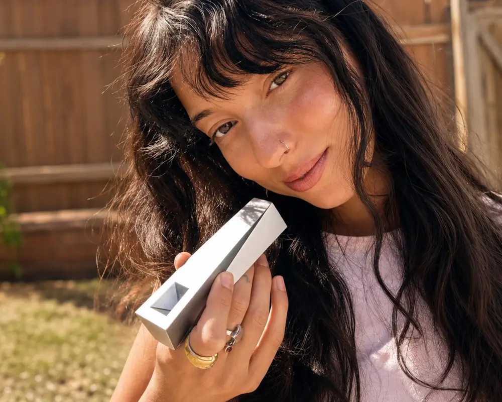 A young woman outdoors holding a silver Tempest pipe with minimalist geometric design