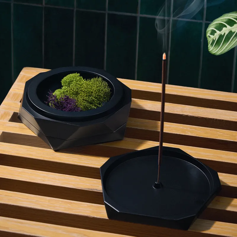 Vesta ashtray in black filled with green and purple moss next to its lid holding a lit incense stick, on a slatted wood surface against dark green tile