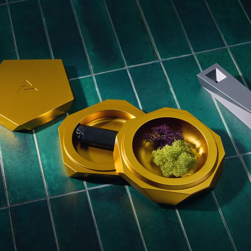 Vesta magnetic ashtray in gold with lid removed, twin compartments holding a black lighter and green and purple moss, silver Tempest pipe resting on dark green tile