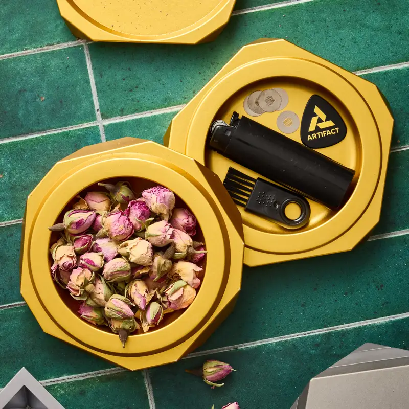 Open gold Vesta container holding dried rosebuds and Artifact accessories on green tile surface