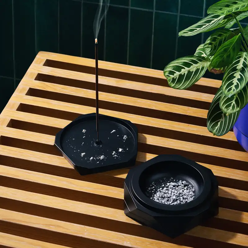 Black Vesta incense holder and ashtray set on slatted wood surface with tropical plant backdrop