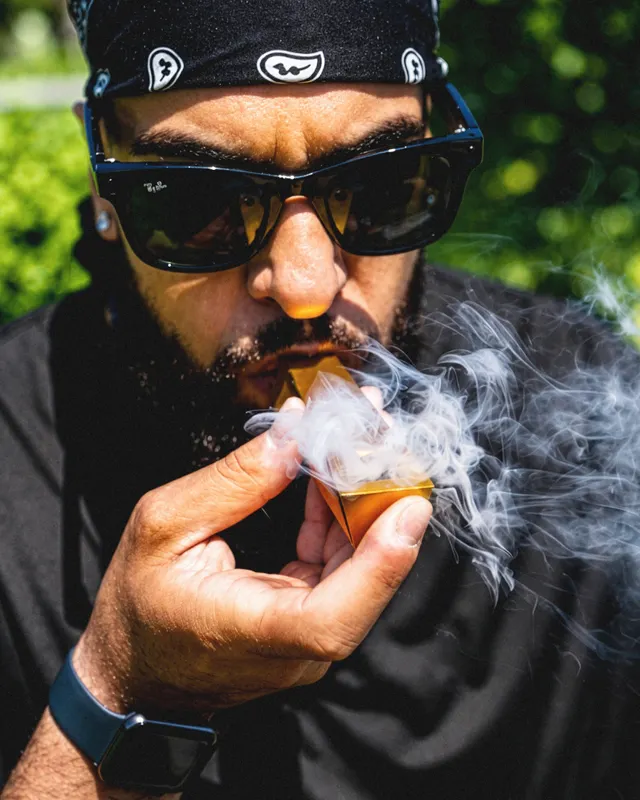 Man taking a hit from a gold Tempest pipe outdoors with visible smoke and minimalist aluminum design