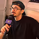 Jeune homme portant des lunettes et une casquette noire, tenant un microphone violet, semblant parler en interview.
