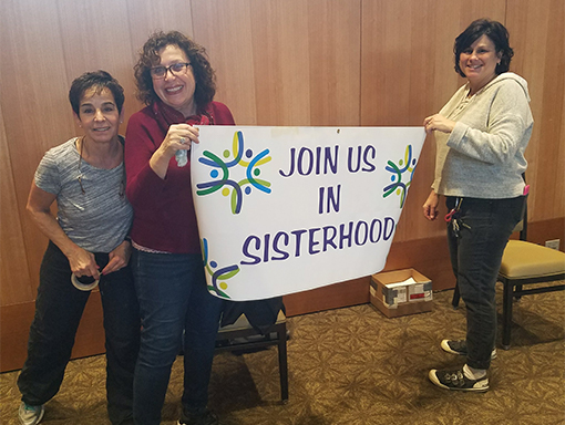 Three smiling women holding a banner that reads, "Join us in sisterhood." 