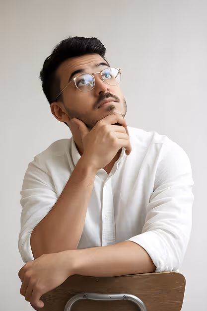 Young man with glasses in a white shirt sitting on a chair, resting chin on hand and looking thoughtfully upward.