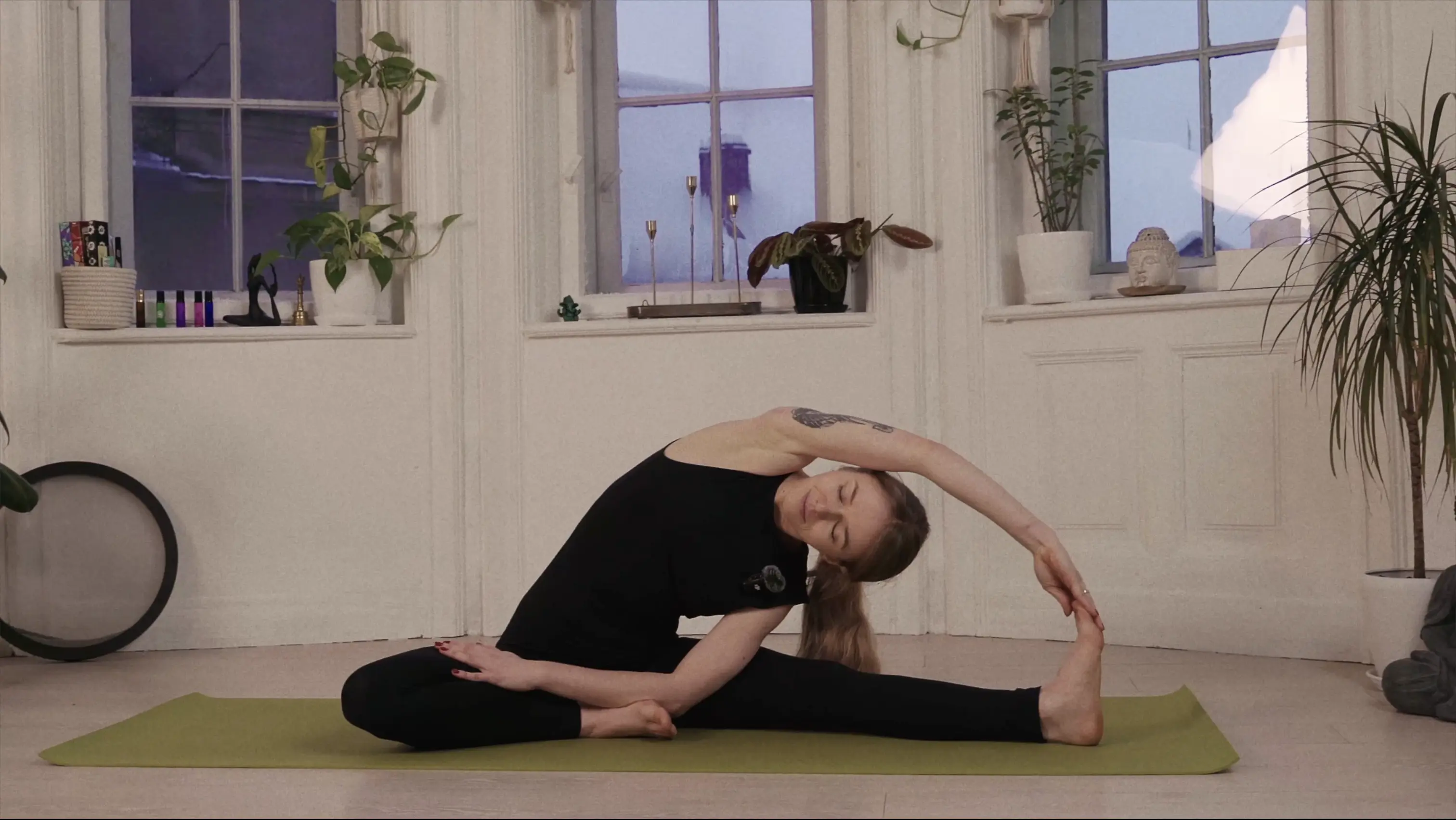 Woman in black athletic wear performing a seated side stretch yoga pose on a green mat in a bright room with potted plants and windows.