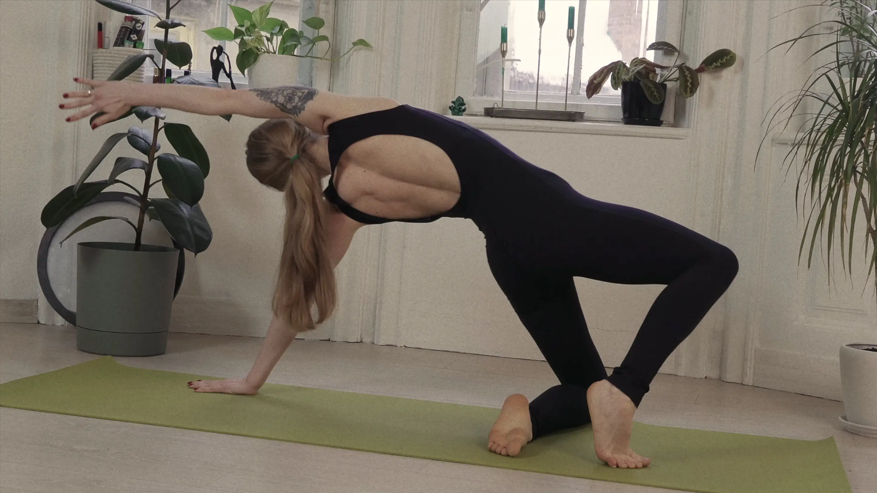 Woman in black leggings and sleeveless top performing side stretch yoga pose on a green mat indoors.