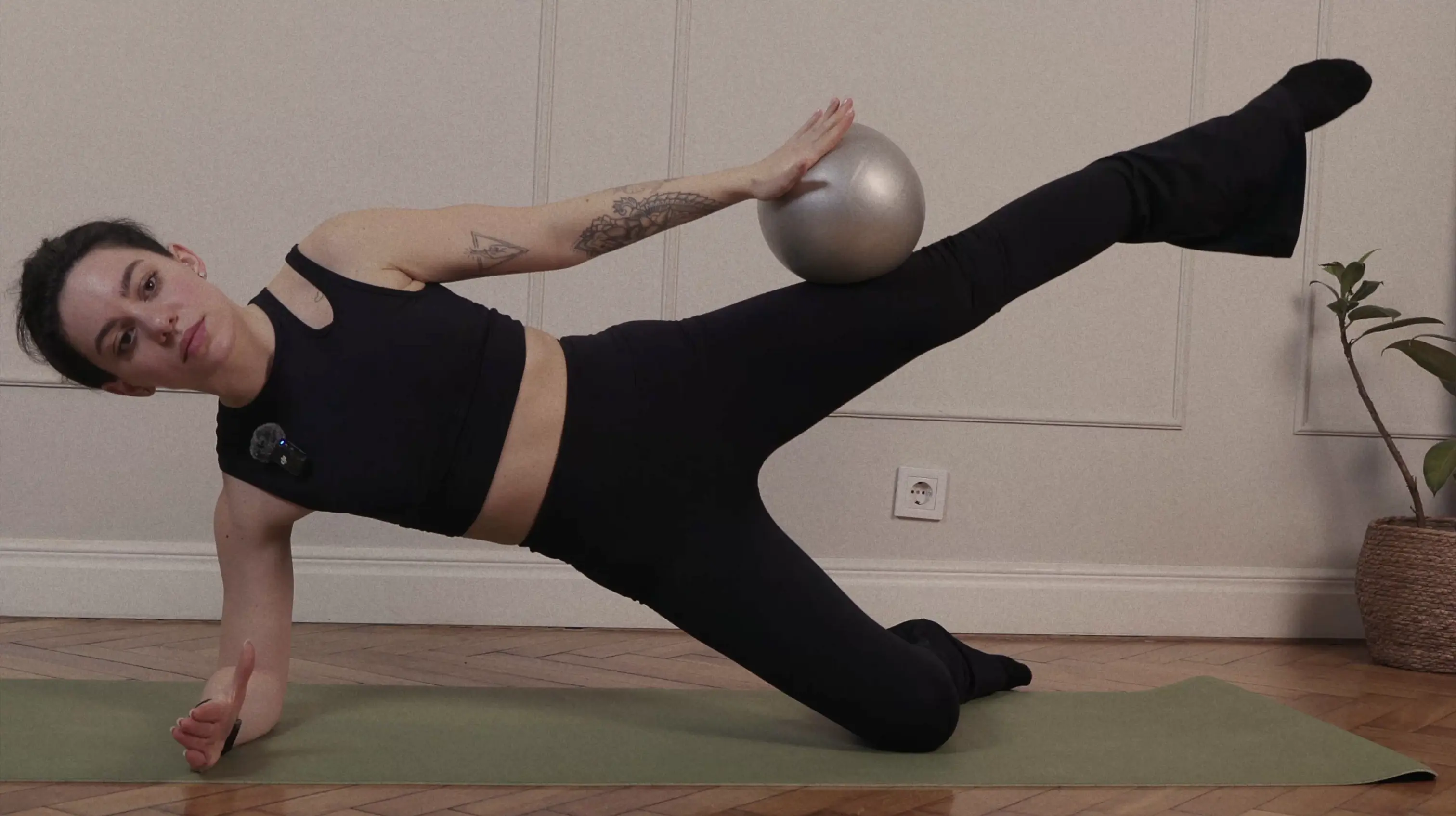 Woman in black workout clothes balancing on one arm and knee on a yoga mat while holding a silver exercise ball on her raised leg.