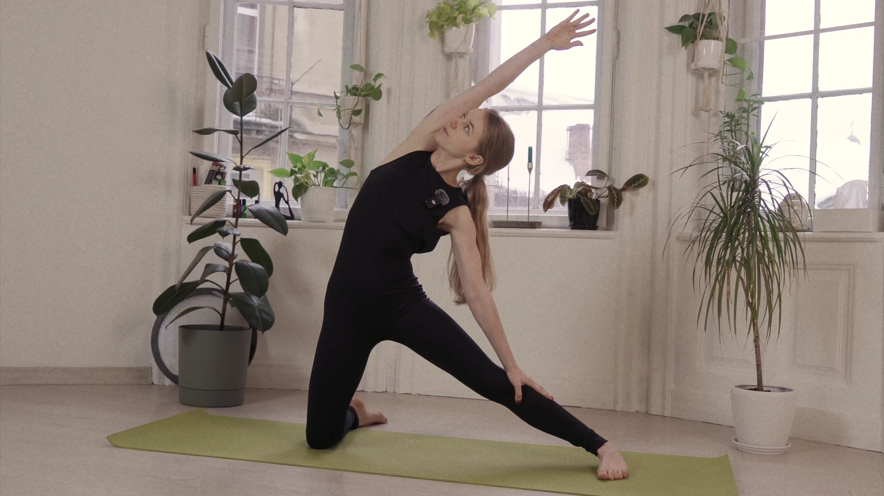 Woman in black workout outfit performing a side stretch yoga pose on a green mat in a bright room with large windows and indoor plants.