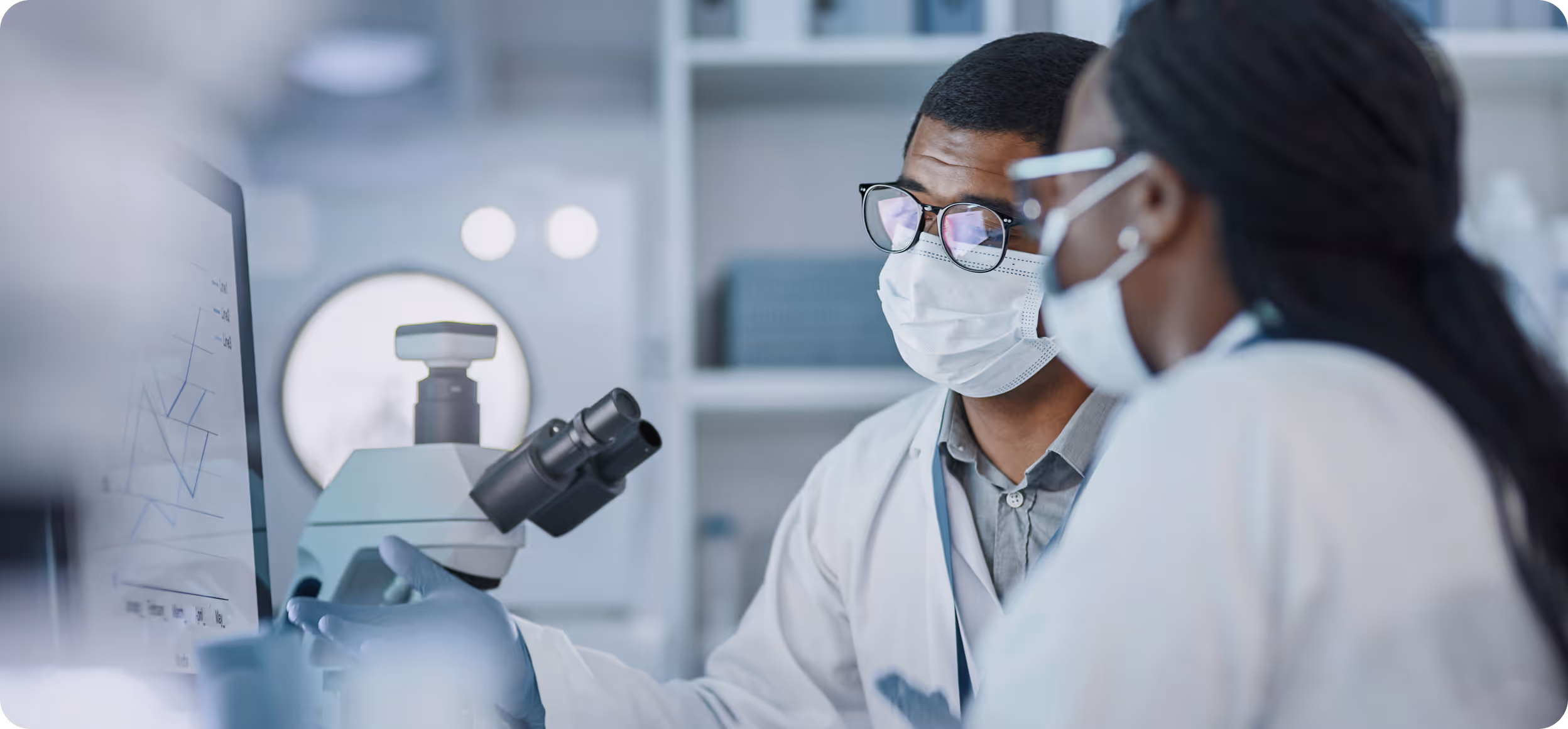 Two scientists in masks discussing data on a computer screen beside a microscope in a lab.