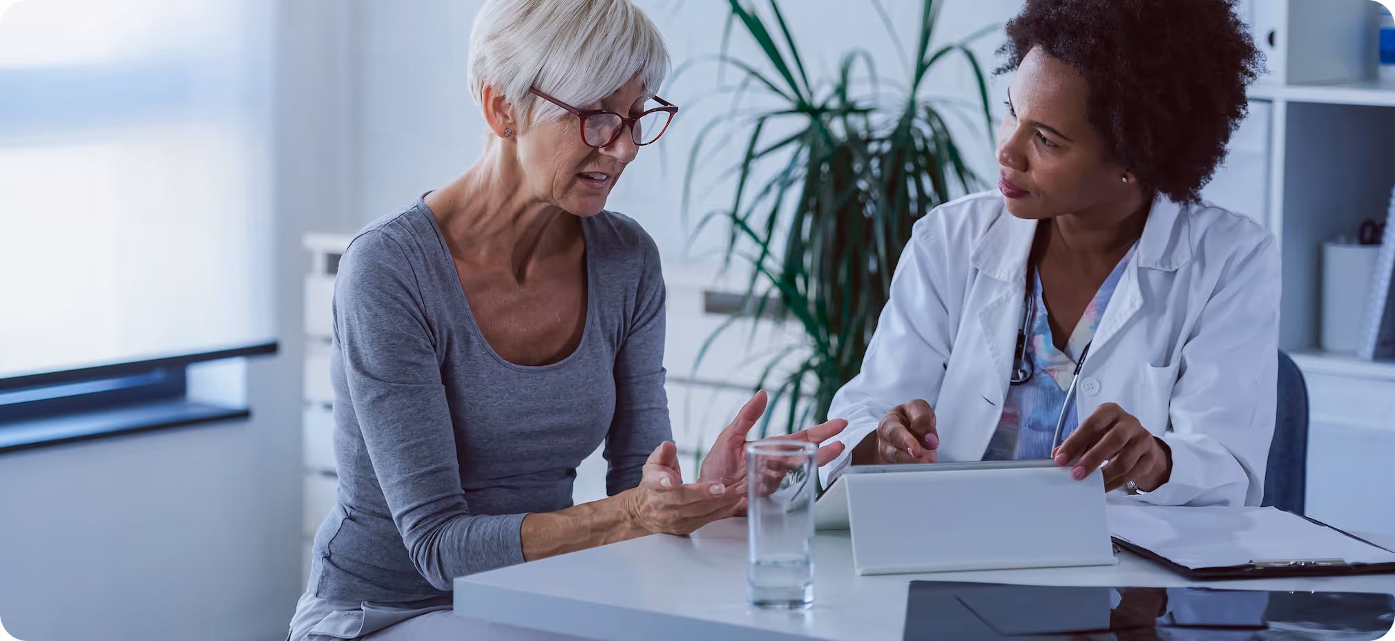 Older woman discussing a clinical diagnosis with a female doctor who is reviewing a tablet in a medical office.