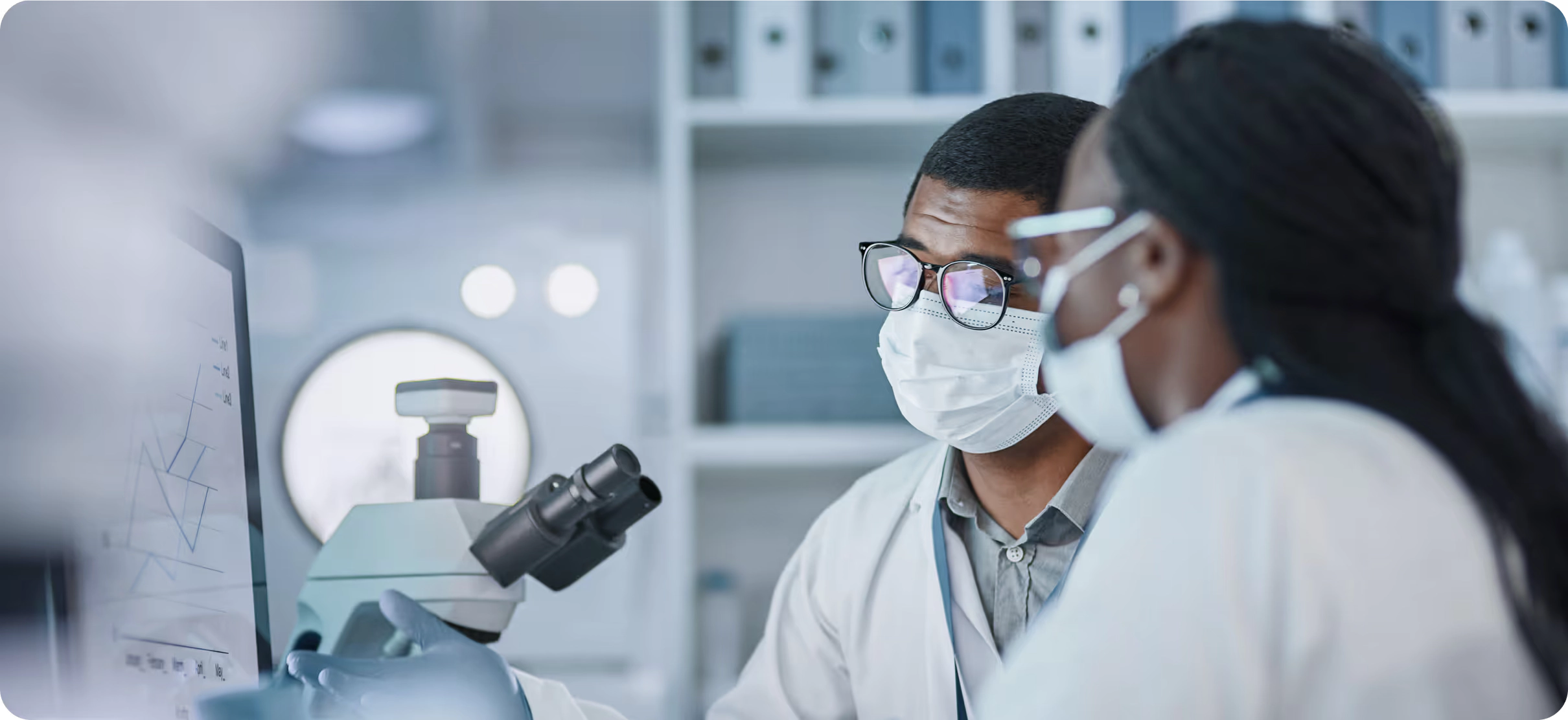 Two researchers in lab coats discussing data on a screen beside a microscope.