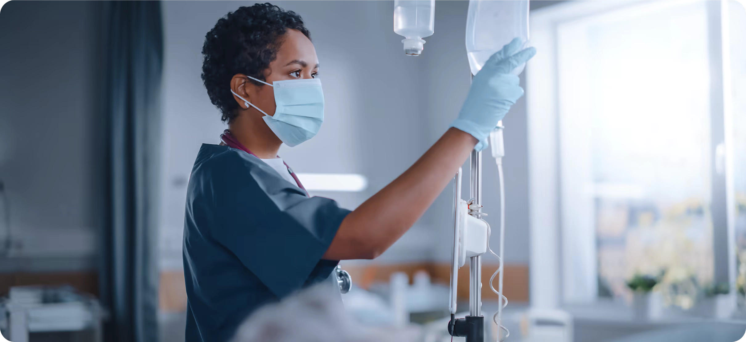 Healthcare worker wearing mask and gloves adjusting an IV drip in a hospital room.