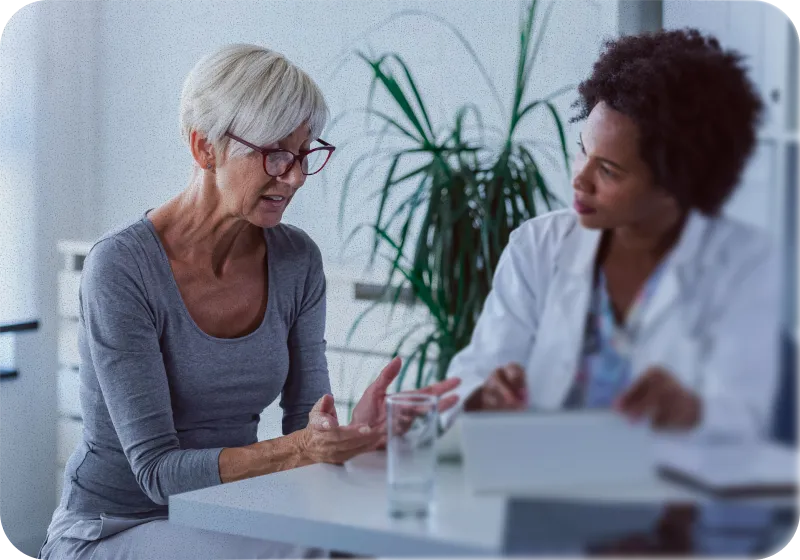 Older woman discussing a clinical diagnosis with a female doctor who is reviewing a tablet in a medical office.