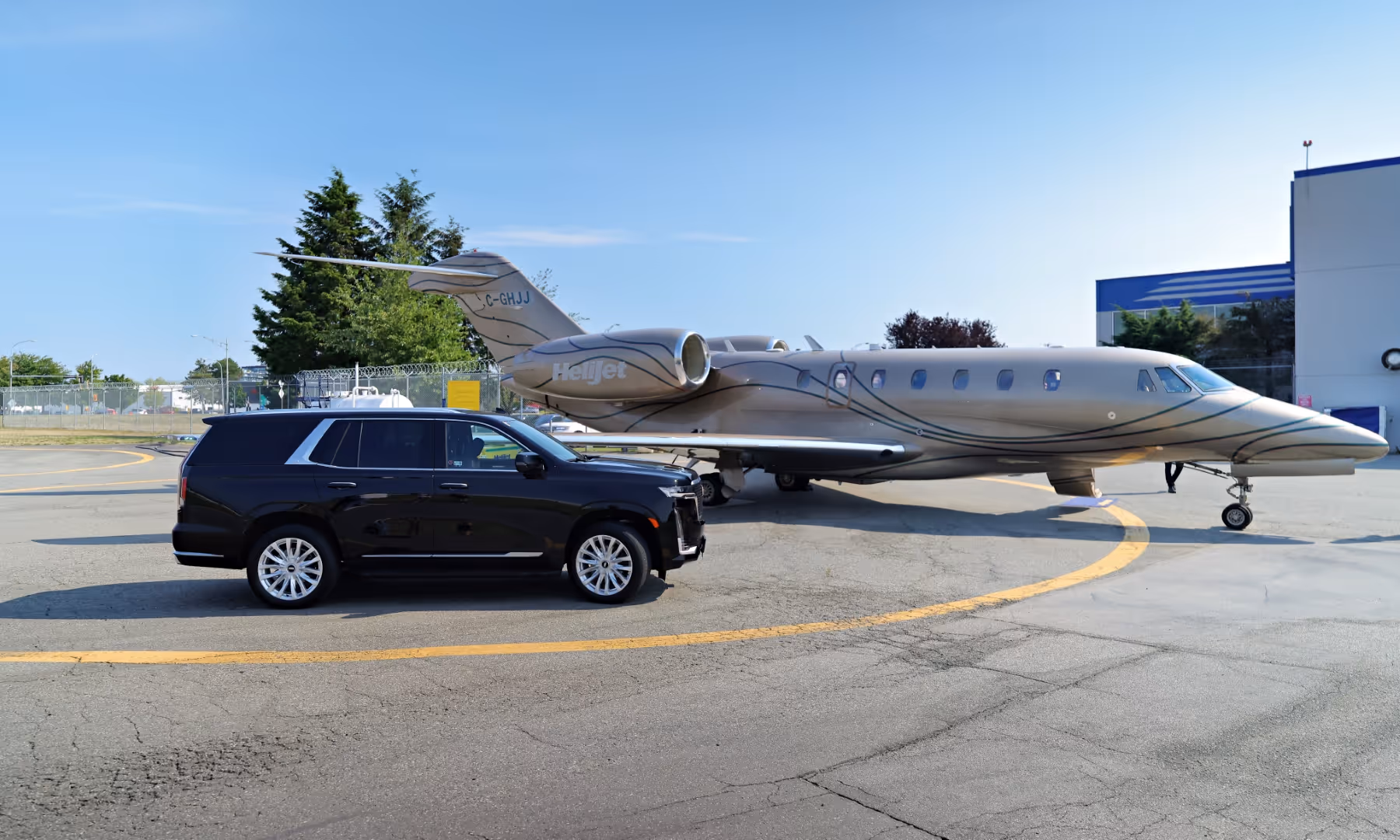 Black luxury Cadillac Escalade SUV parked next to a beige private jet with Helijet branding on a sunny airport tarmac.