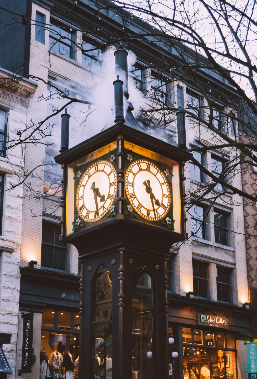 Historic Gastown steam clock with glowing clock faces and steam rising at dusk in front of brick buildings.