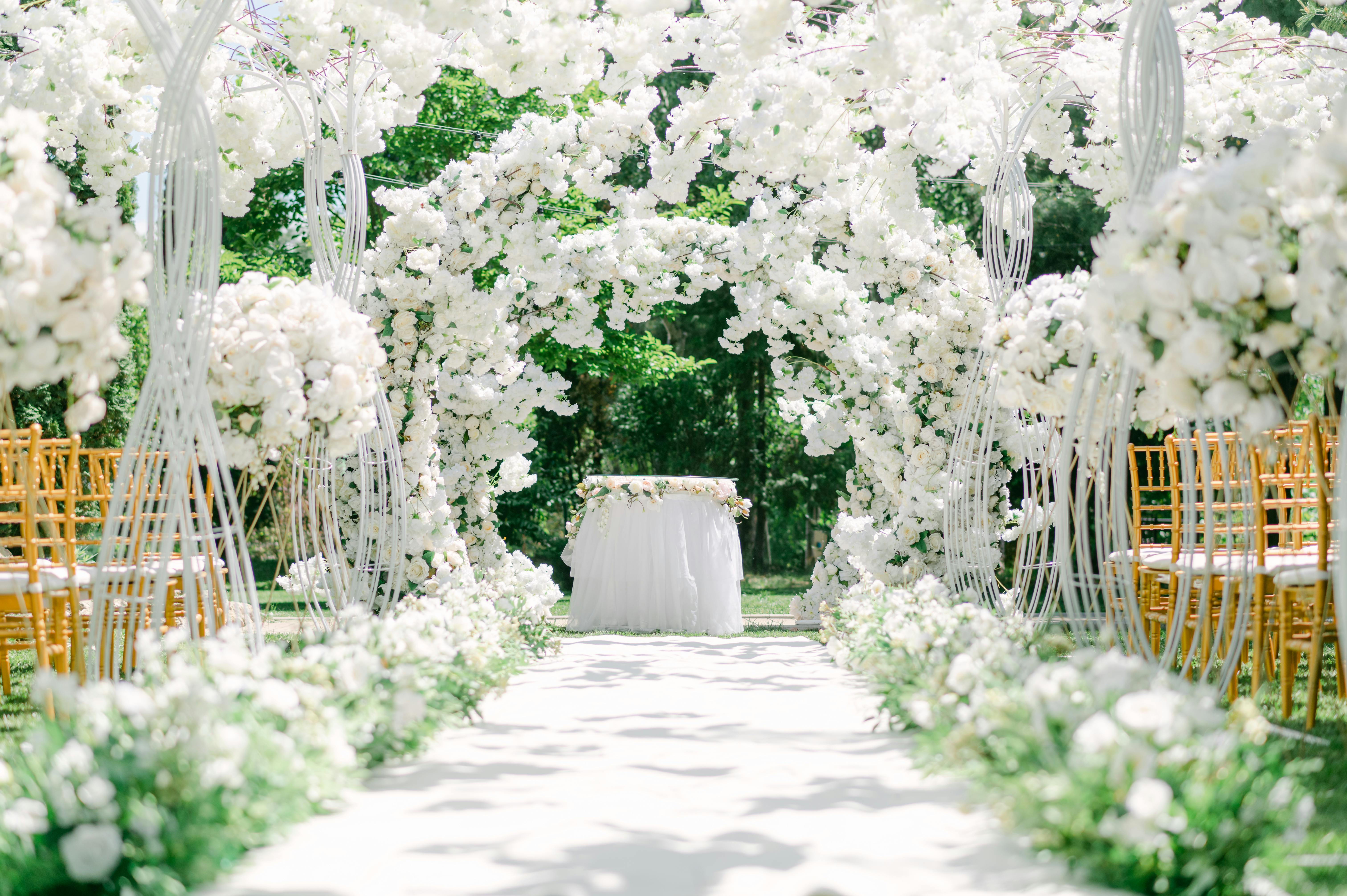 Outdoor wedding aisle lined with white flowers, golden chairs, and white floral arches under sunlight. Credit Soulseeker, Pexels