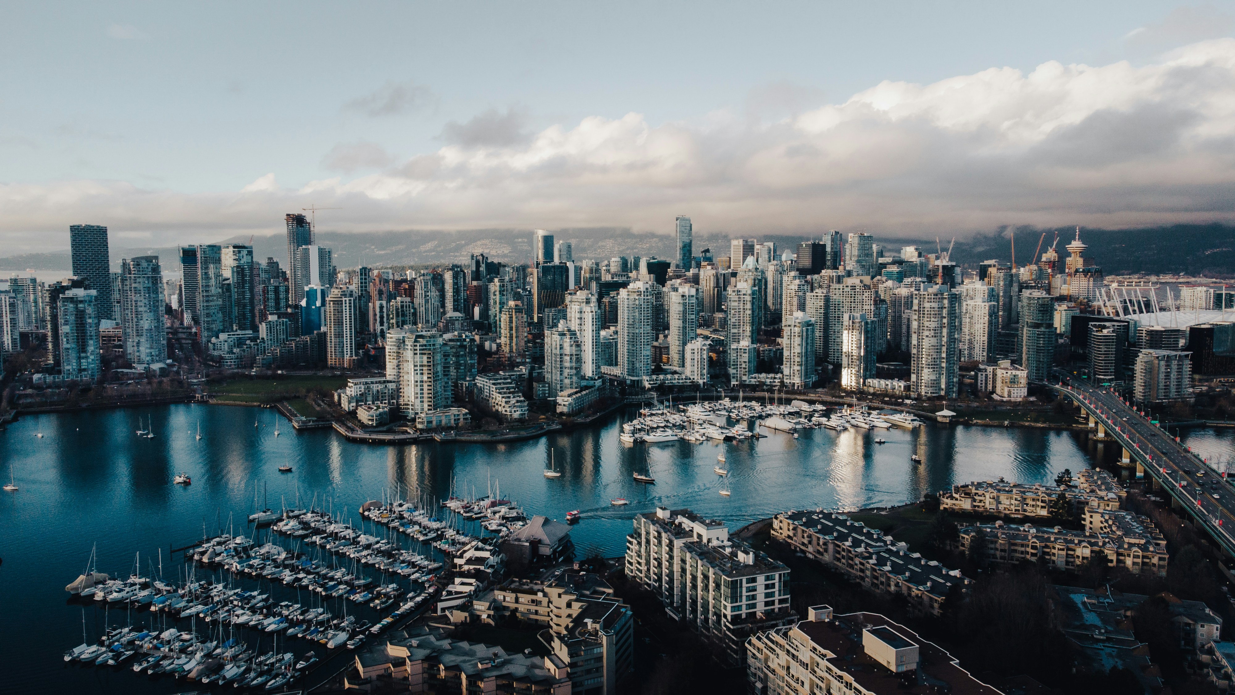 Aerial view of a Vancouver's False Creek city skyline with high-rise buildings, a marina filled with boats, and a bridge over water under a cloudy sky. Credit Matt Wang via Unsplash