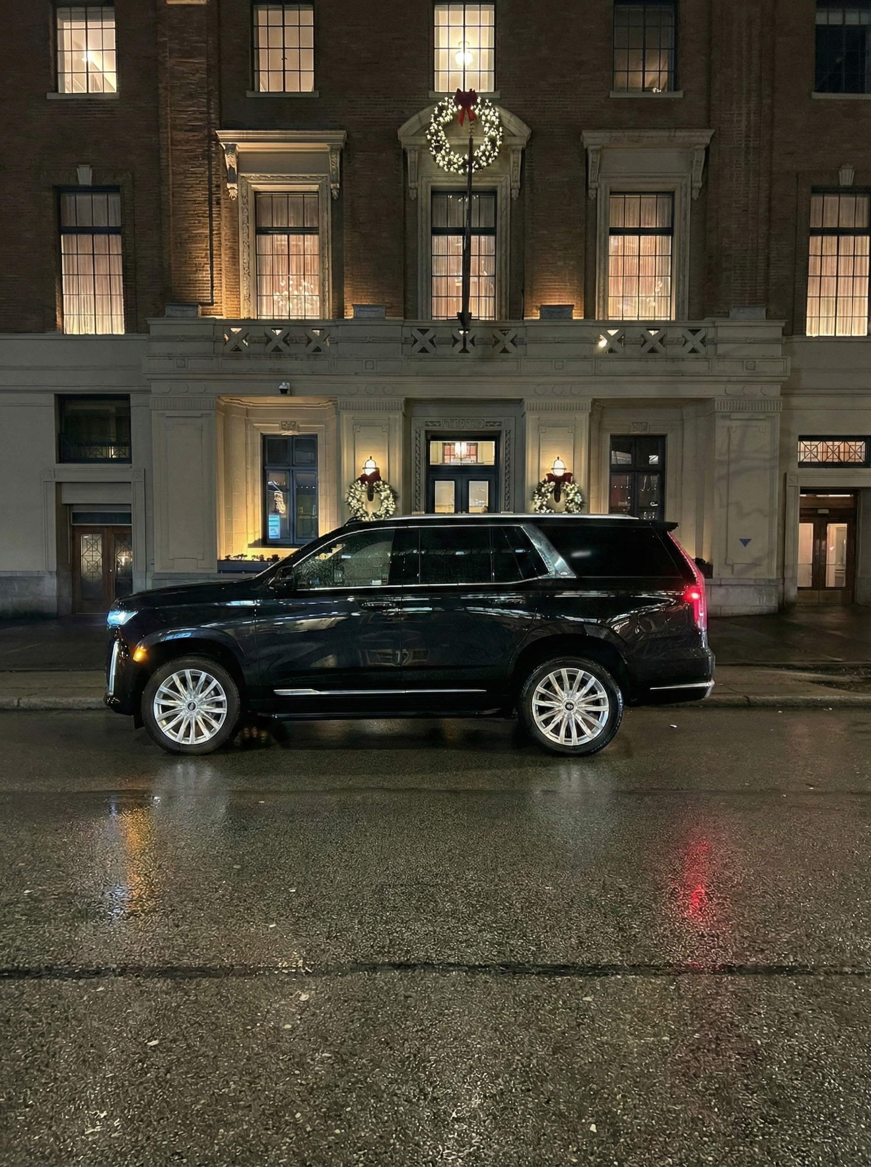 Black Cadillac Escalade luxury SUV parked on a wet street at night in front of the Vancouver Club, a historic building decorated with illuminated holiday wreaths.