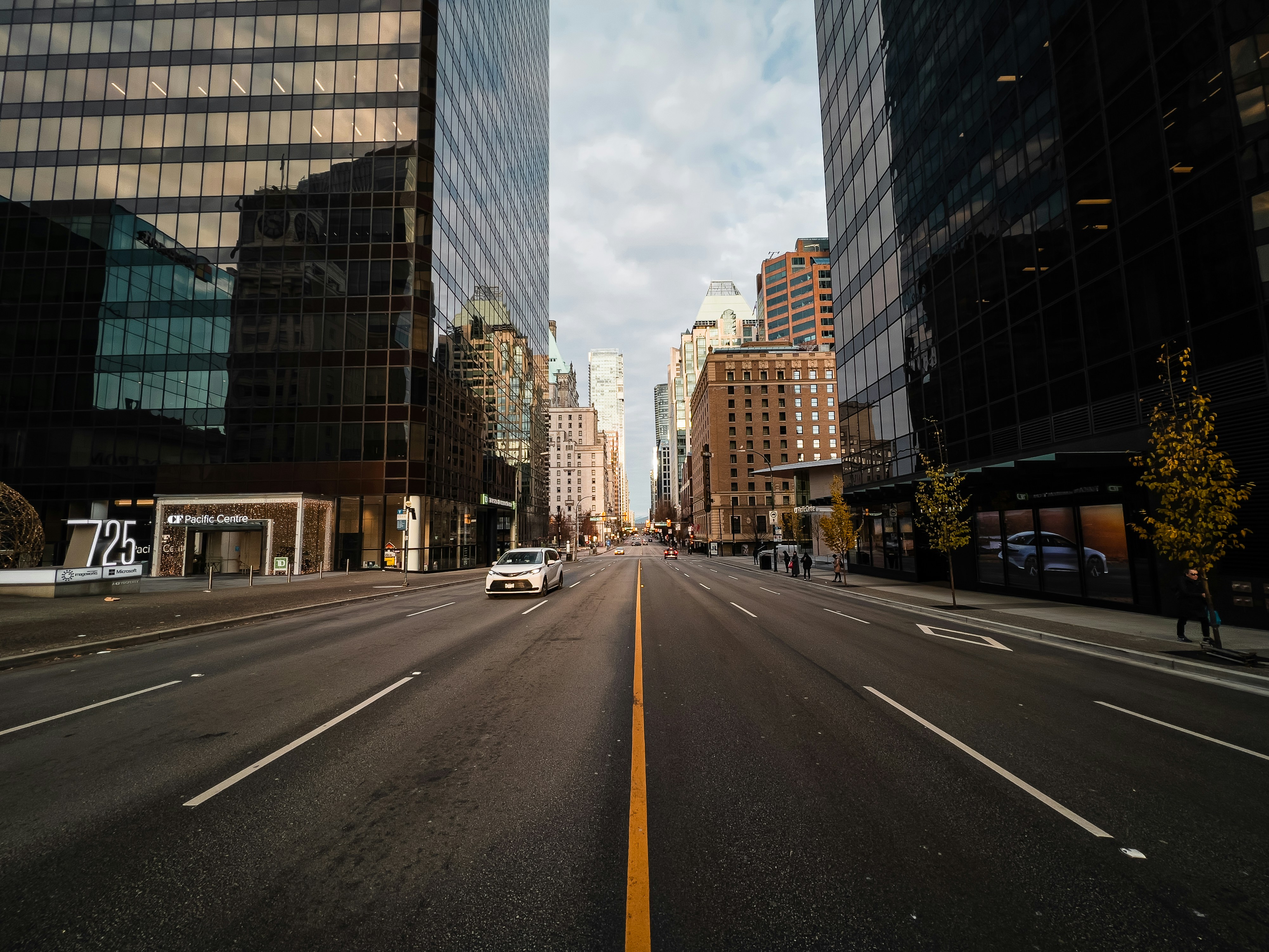 Wide West Georgia street in Vancouver lined with tall buildings and a few pedestrians, with a cloudy sky above. Credit to Aditya Chinchure via Unsplash