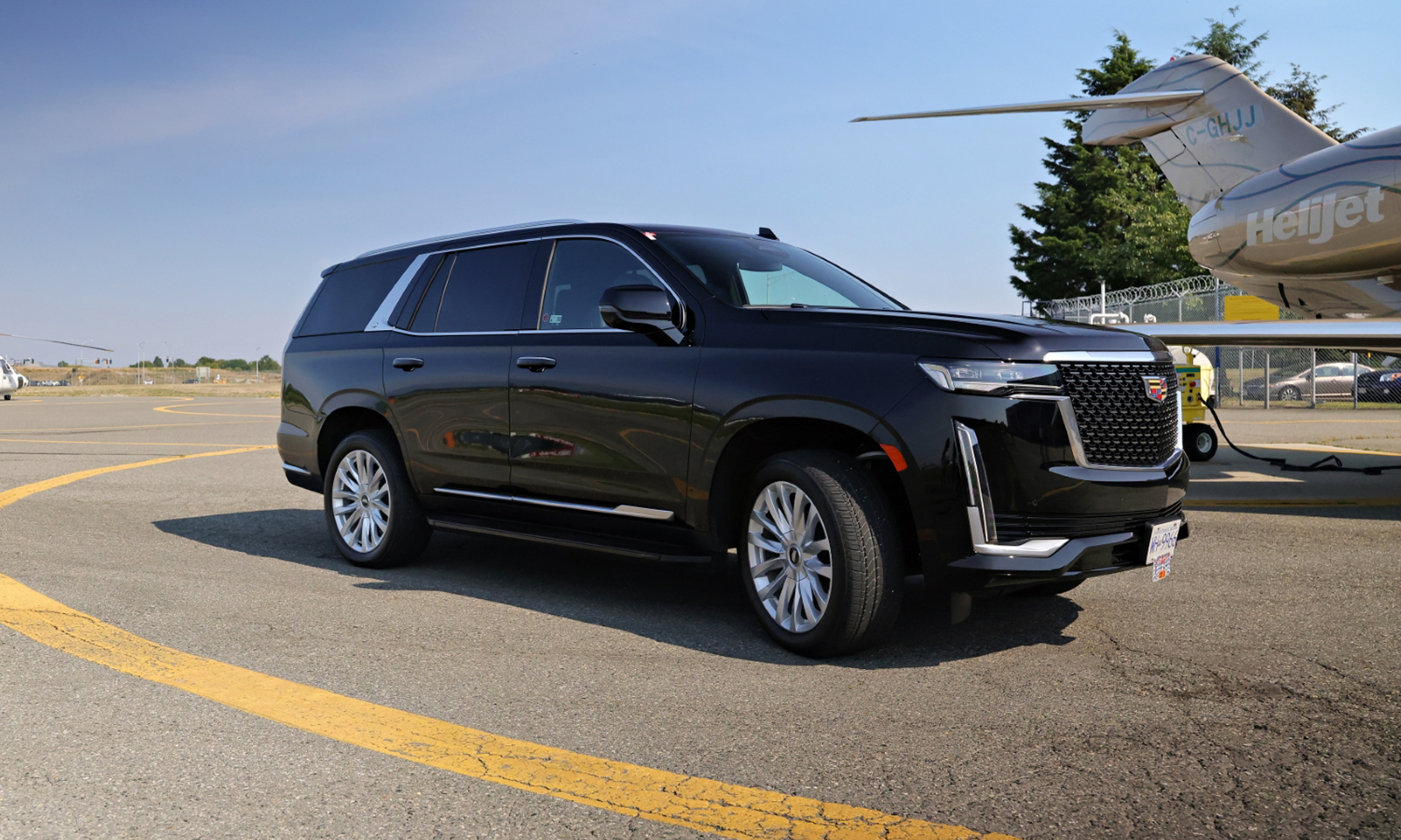 Black Cadillac Escalade SUV parked on airport tarmac near a small aircraft with HellJet branding on the tail.