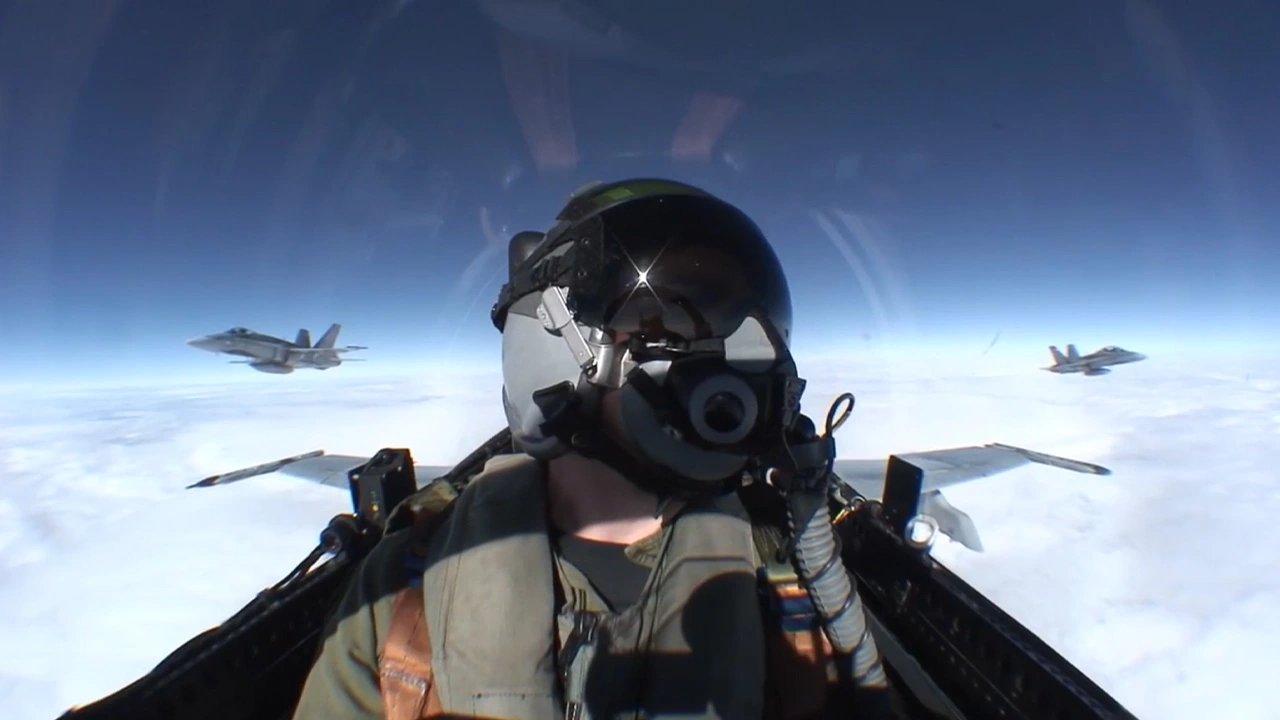 Pilot wearing helmet and oxygen mask inside a fighter jet cockpit flying above clouds with two other fighter jets visible in formation.
