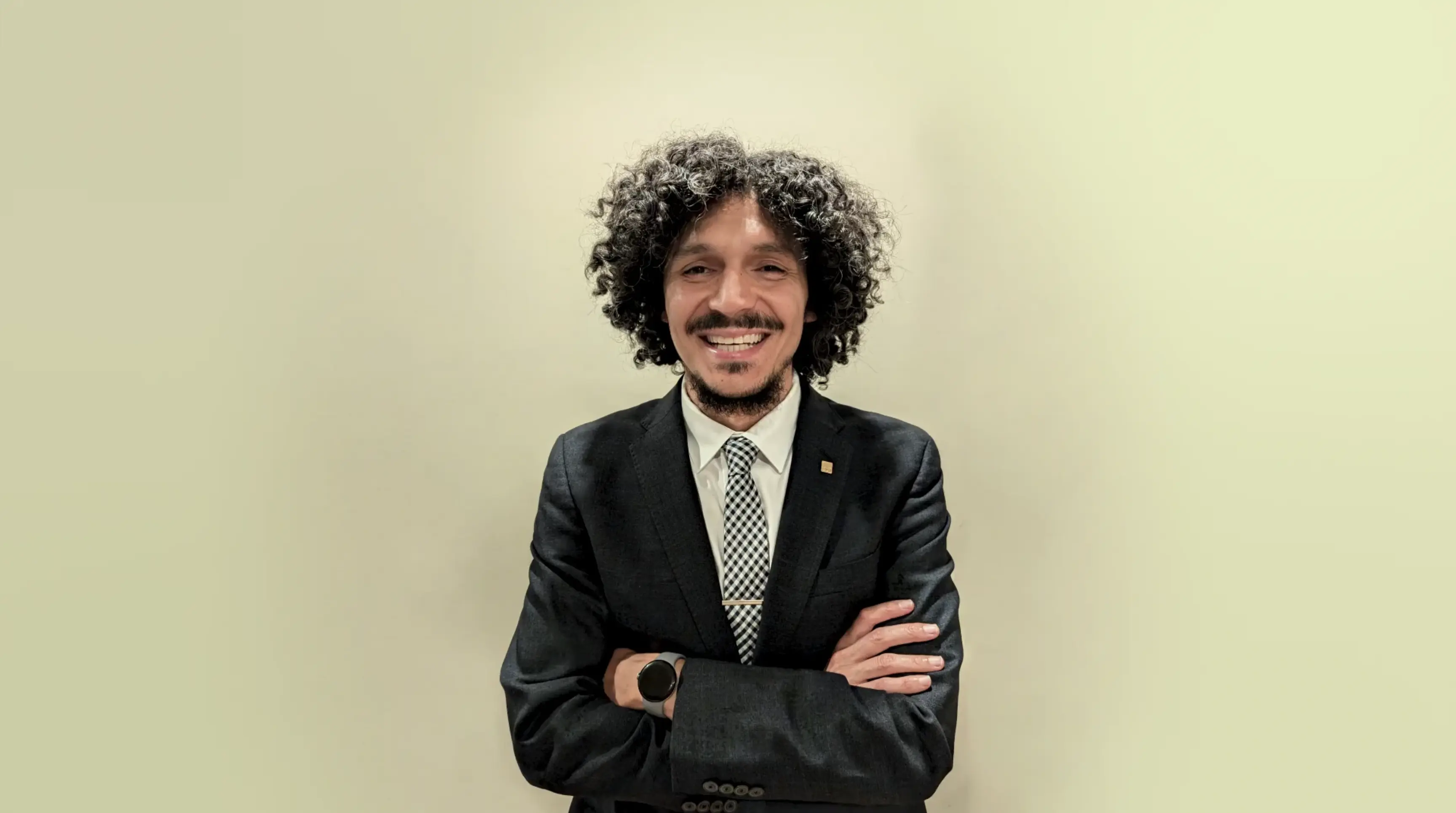 Smiling man with curly hair in a black suit and checkered tie standing with arms crossed against a beige background.