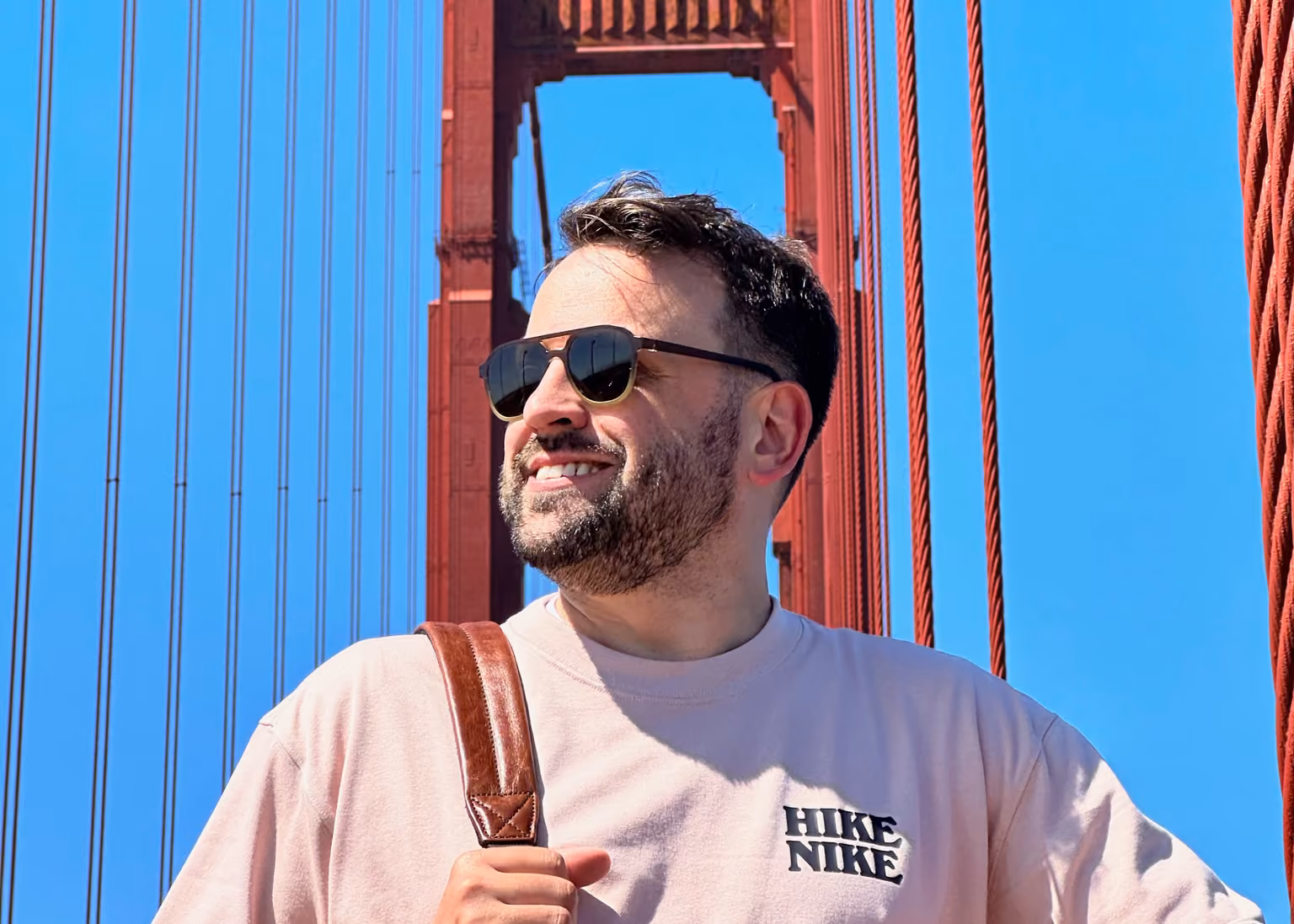 Smiling man with sunglasses and beard on the Golden Gate Bridge wearing a light shirt and backpack.