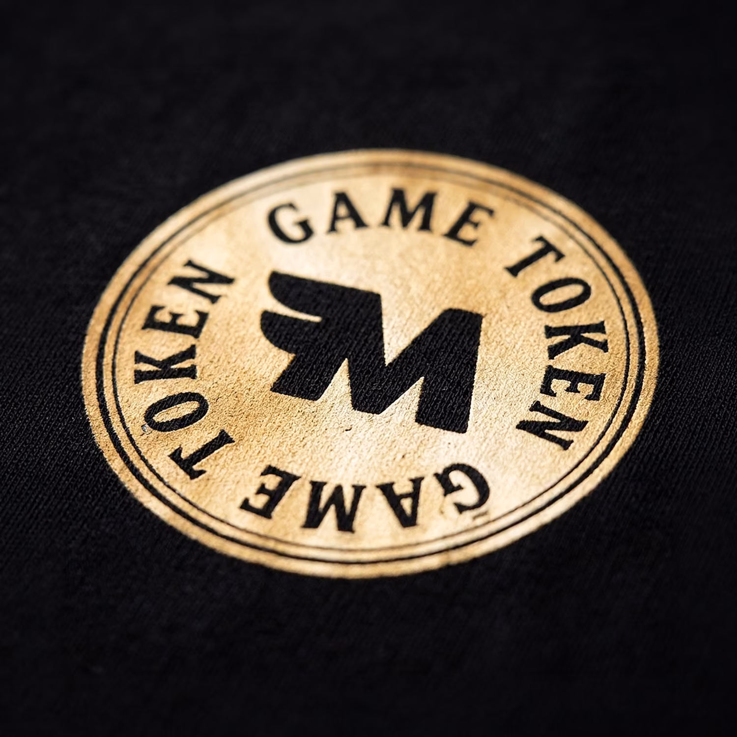 Round worn game token with black winged 'M' logo on black fabric background.