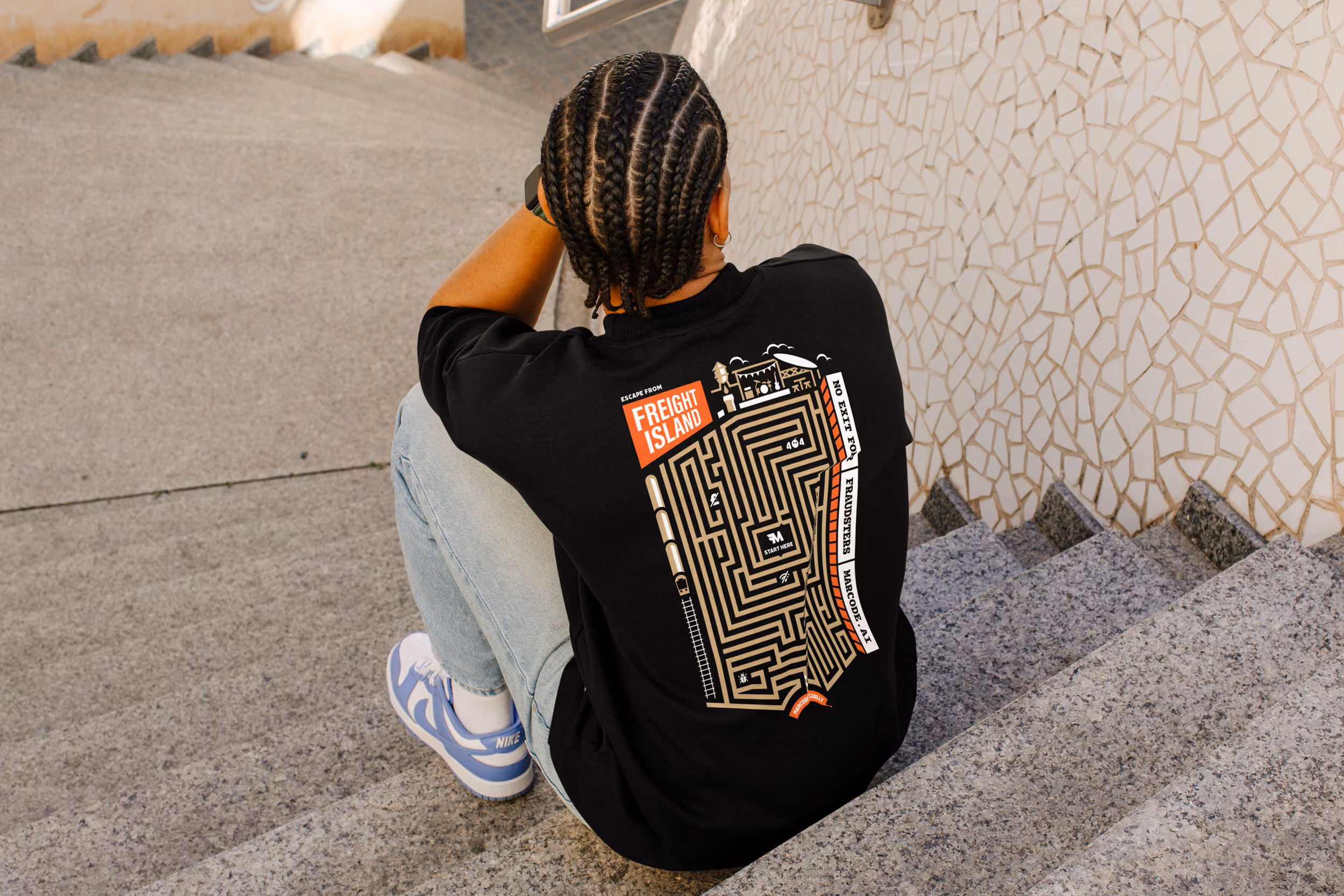 Person with braided hair sits on stone steps wearing black shirt with maze graphic and text on back.