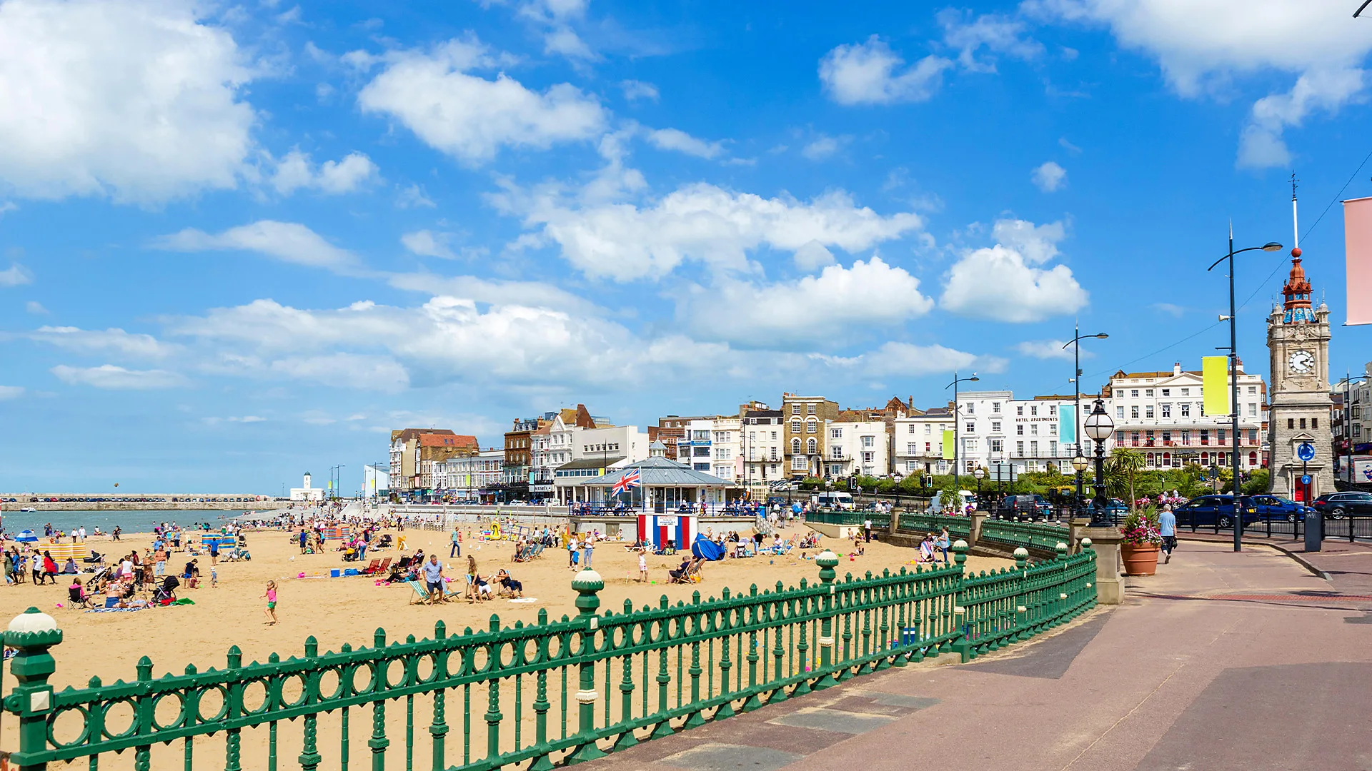 Crowded sandy beach with people, a green railing, seaside buildings, a clock tower, and blue sky with clouds.