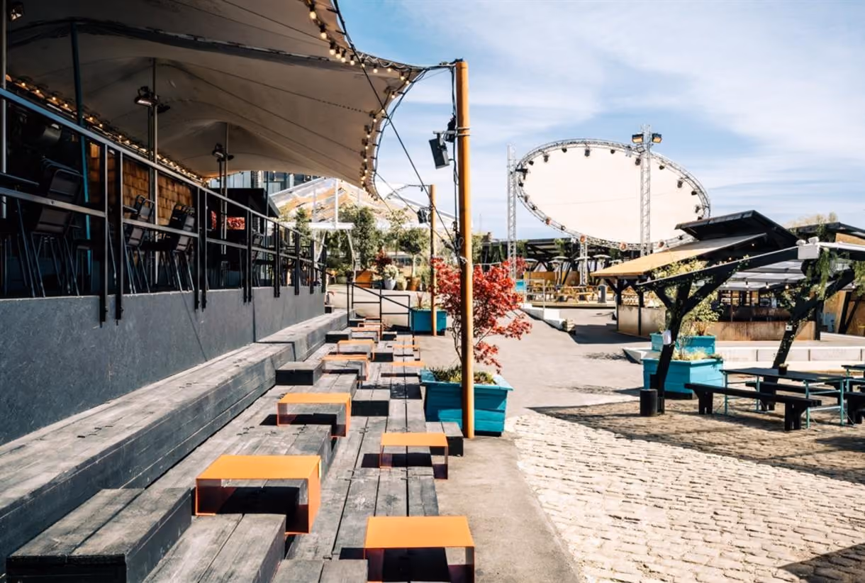 Outdoor seating area with wooden benches, orange tables, planters, and a large white circular stage backdrop.