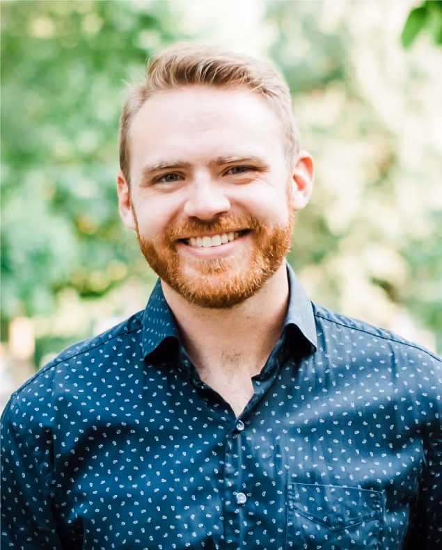 Smiling man with red beard and hair wearing a blue patterned shirt outdoors with greenery background.