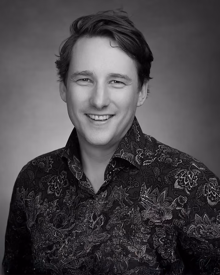 Smiling man with short hair wearing a patterned shirt against a plain background in black and white.