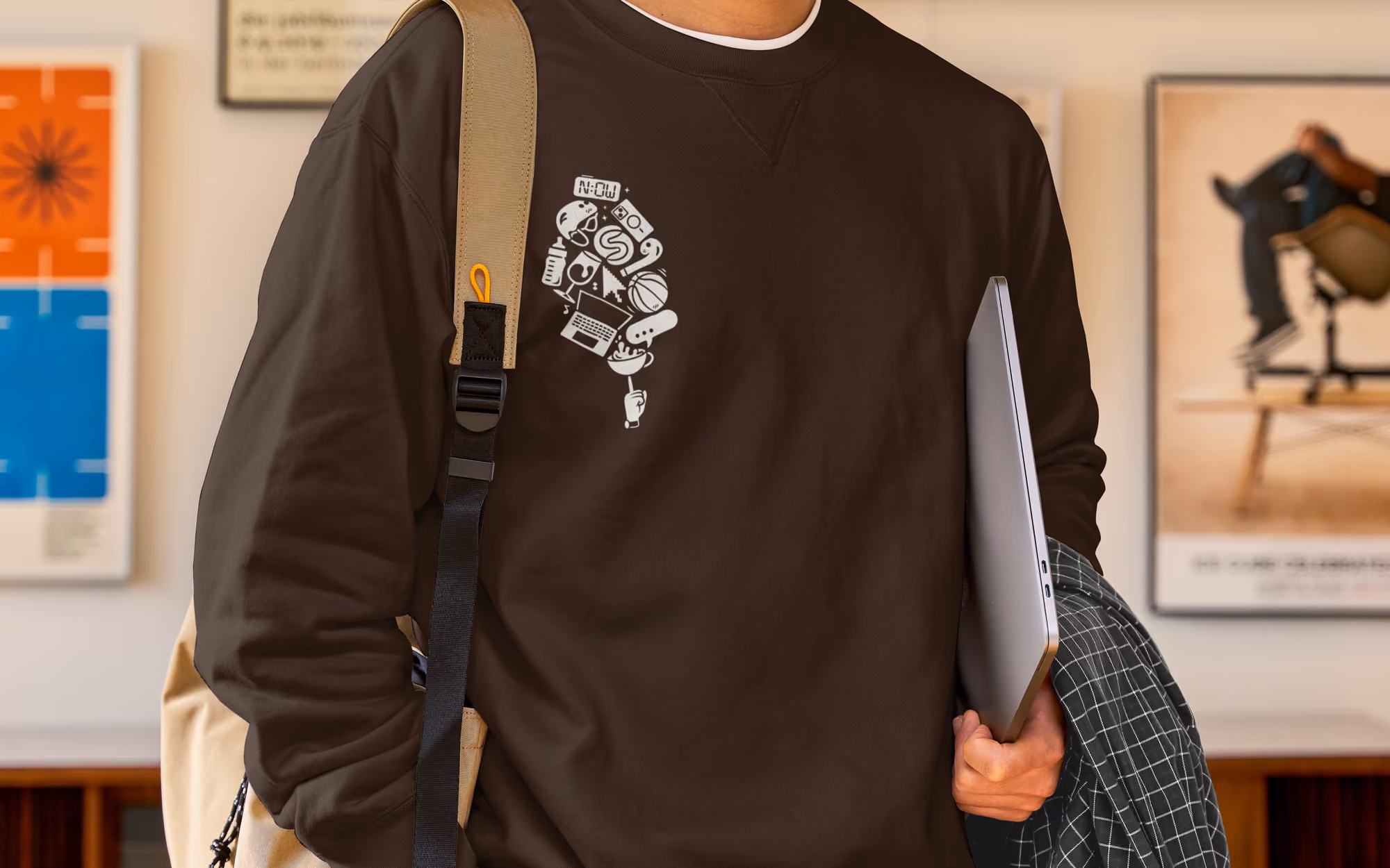 Person wearing brown sweatshirt with white graphic of various icons and holding a laptop under arm.