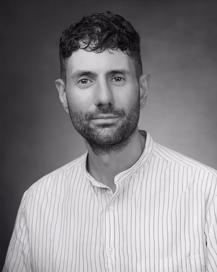 Portrait of a man with short curly hair and beard wearing a striped button-up shirt, black and white.