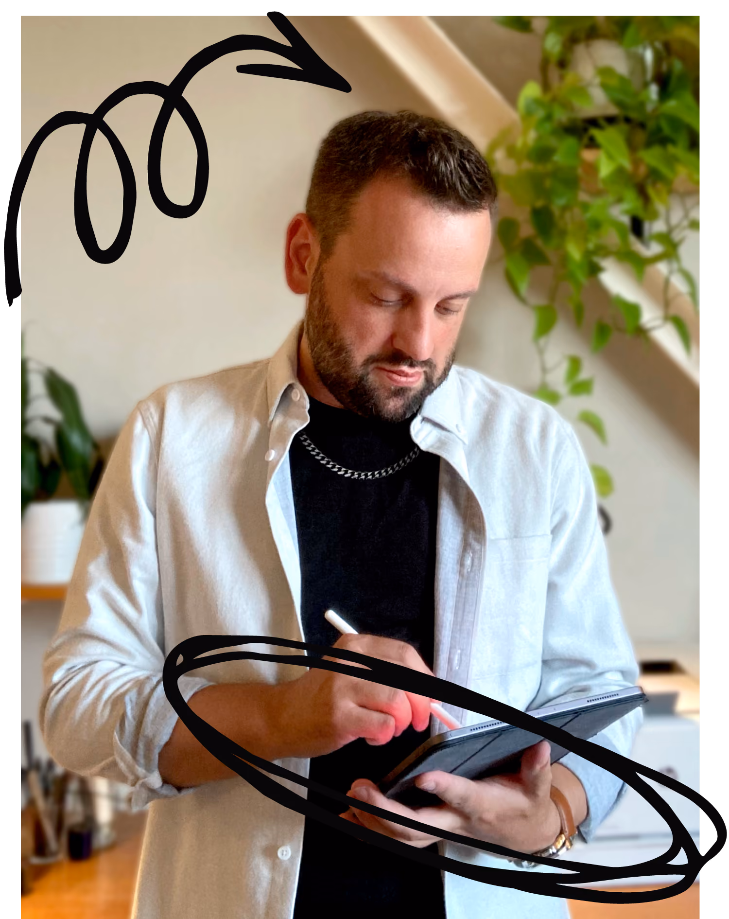 Man with beard using digital pen on tablet with plants in background and white shirt over black shirt.