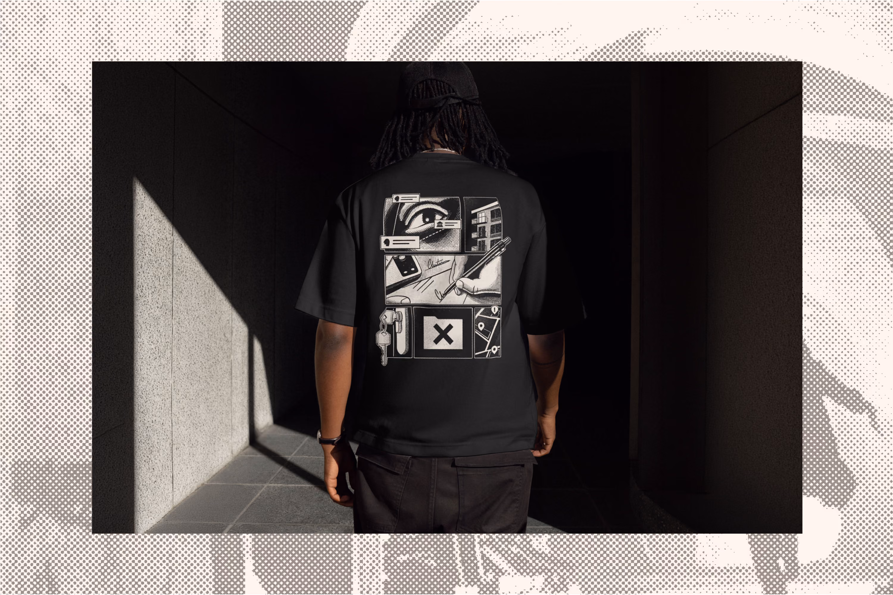 Person wearing a black t-shirt with graphic designs on the back, including an eye, hand holding a stick, keys, and other abstract elements, standing in a shadowed corridor with light on a concrete wall.