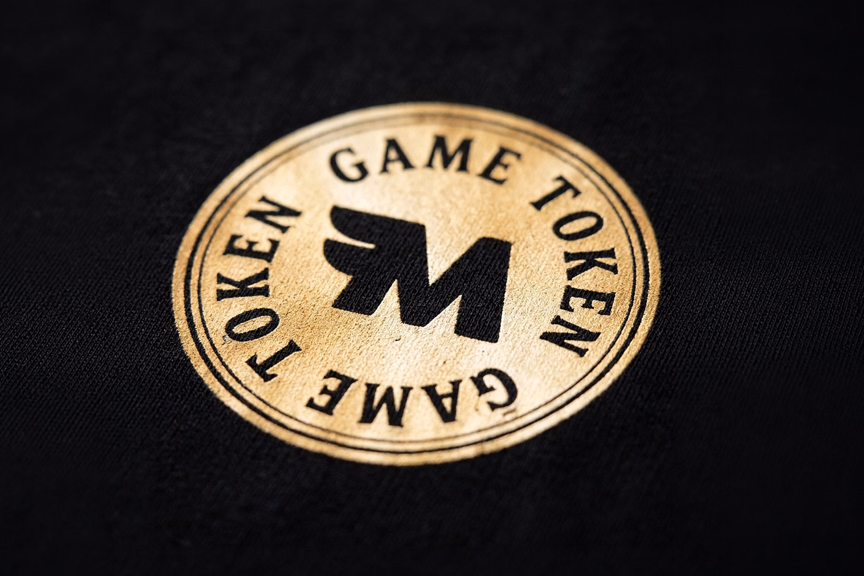 Round gold-colored token with the letters 'M' in the center and the words 'GAME TOKEN' written twice around the edge on a black fabric background.