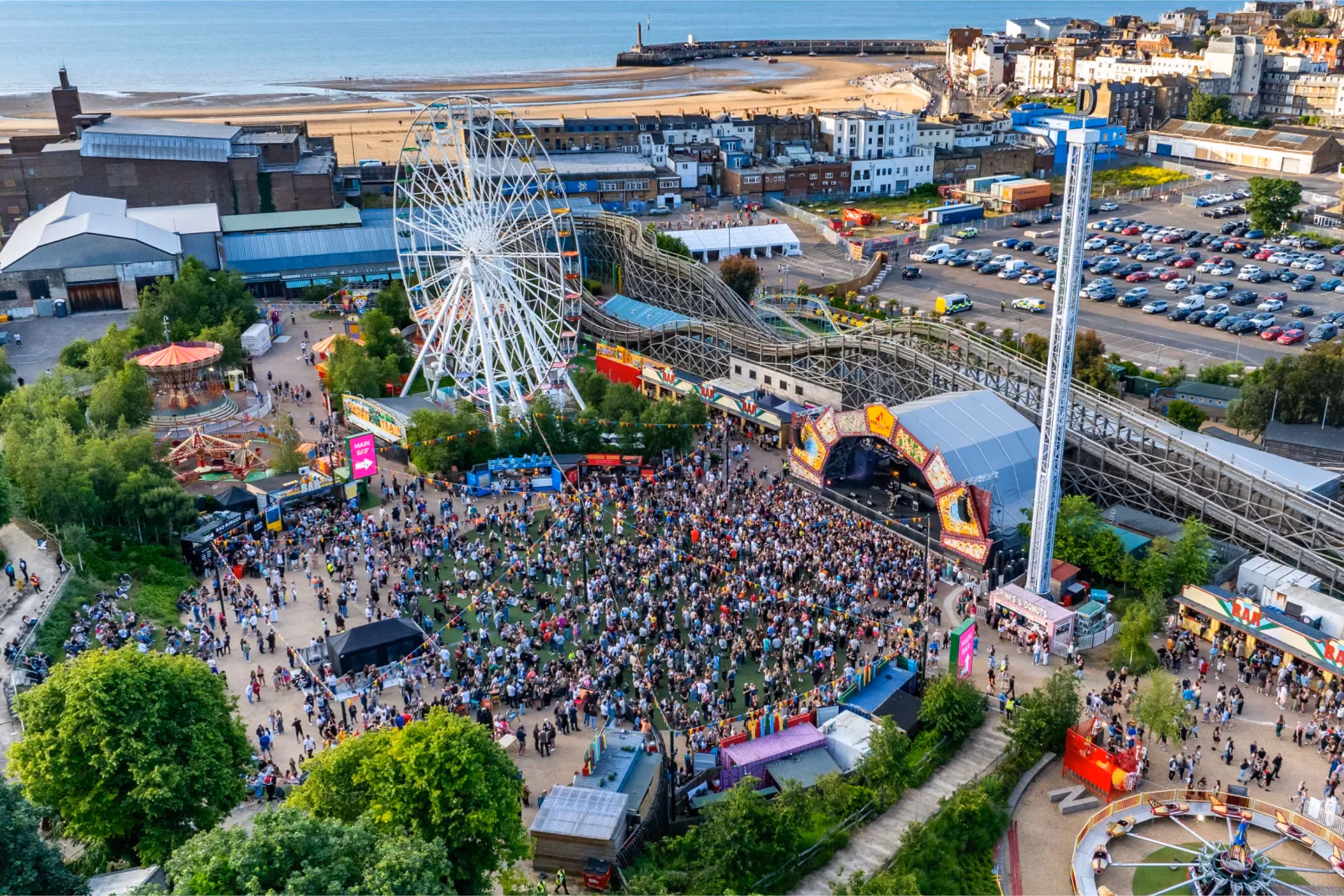 Aerial view of a crowded amusement park with a large Ferris wheel, roller coaster, stage with a performance, and various attractions, with the sea and harbor visible in the background.