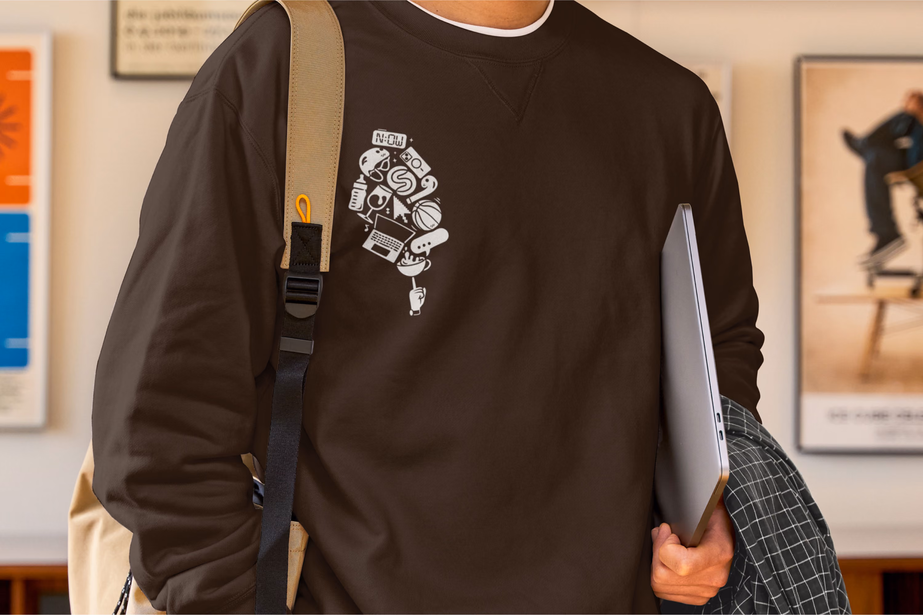 Person wearing a dark brown sweatshirt with a white graphic of a stylized head made of icons such as a laptop, dollar sign, basketball, cassette tape, and drink cup, holding a laptop and a checkered jacket.