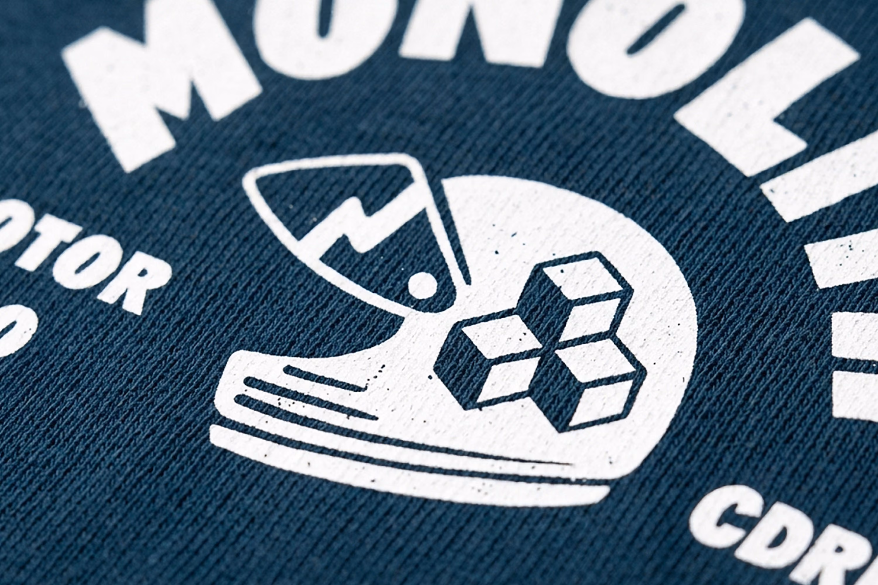 Close-up shot of a navy blue fabric with a white graphic design featuring a stylized helmet with a lightning bolt and geometric shapes, part of the text 'MONOLI' visible above and 'CDR' below.