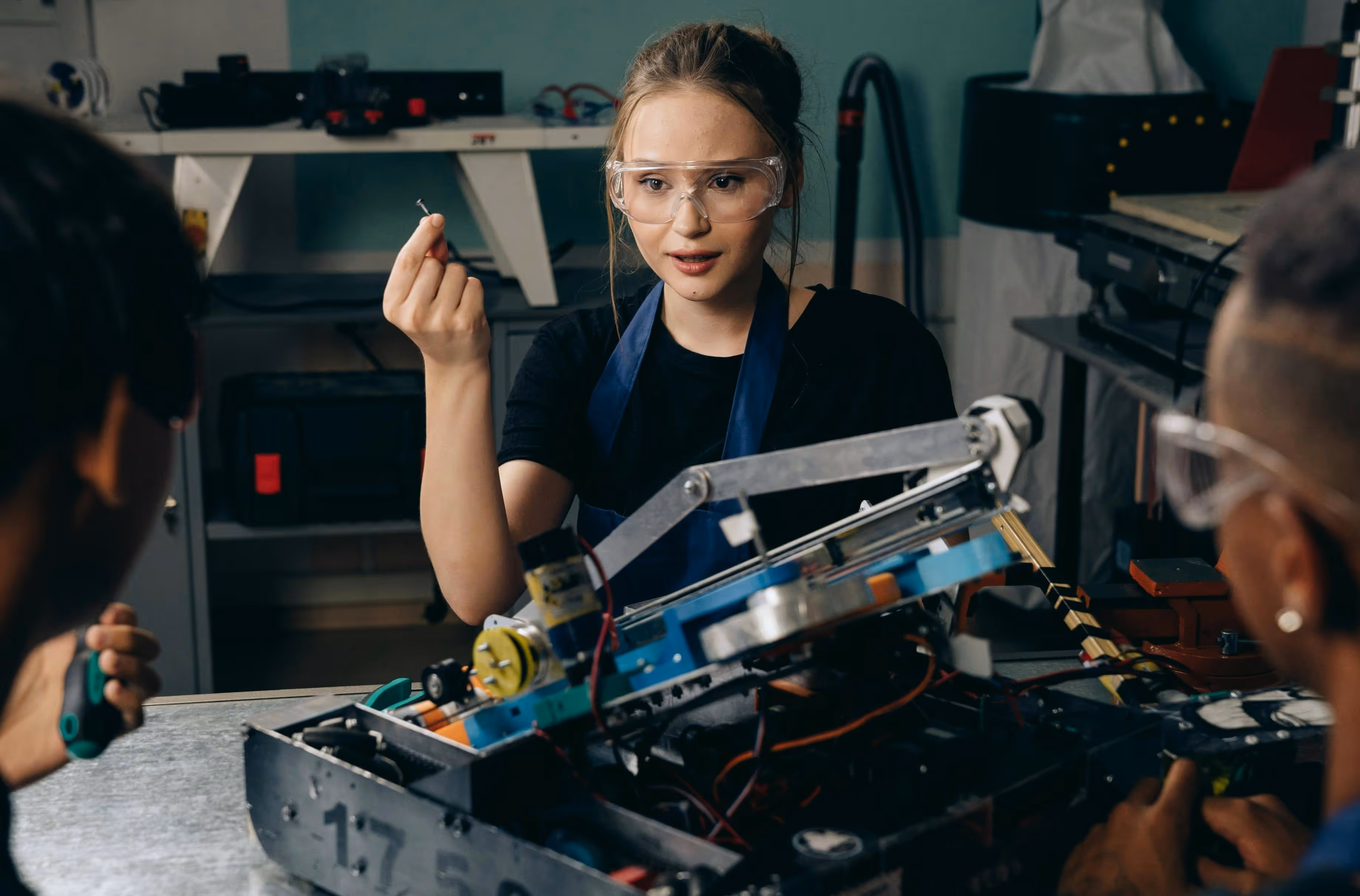 A young woman with safety glasses eagerly holds up a part to a robot that she is building with two other teammates. They are sitting on opposite sides of the table, listening to her speak, with the robot on the table in front of them.