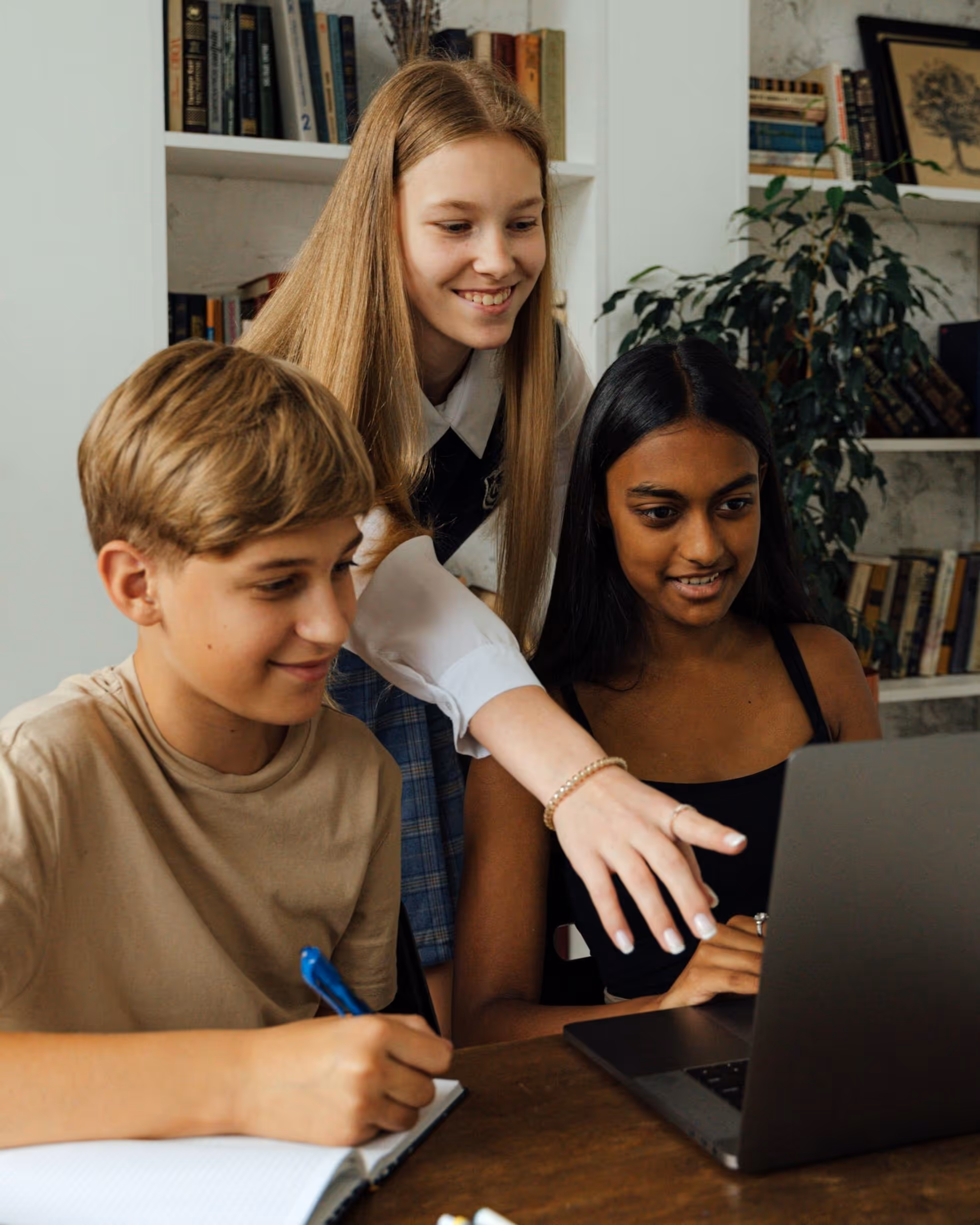 A group of three teenagers doing homework together in front of a laptop. The older girl in the middle is tutoring a younger boy and girl seated at the table. She is standing behind them, pointing something out on the laptop screen, while they take notes.