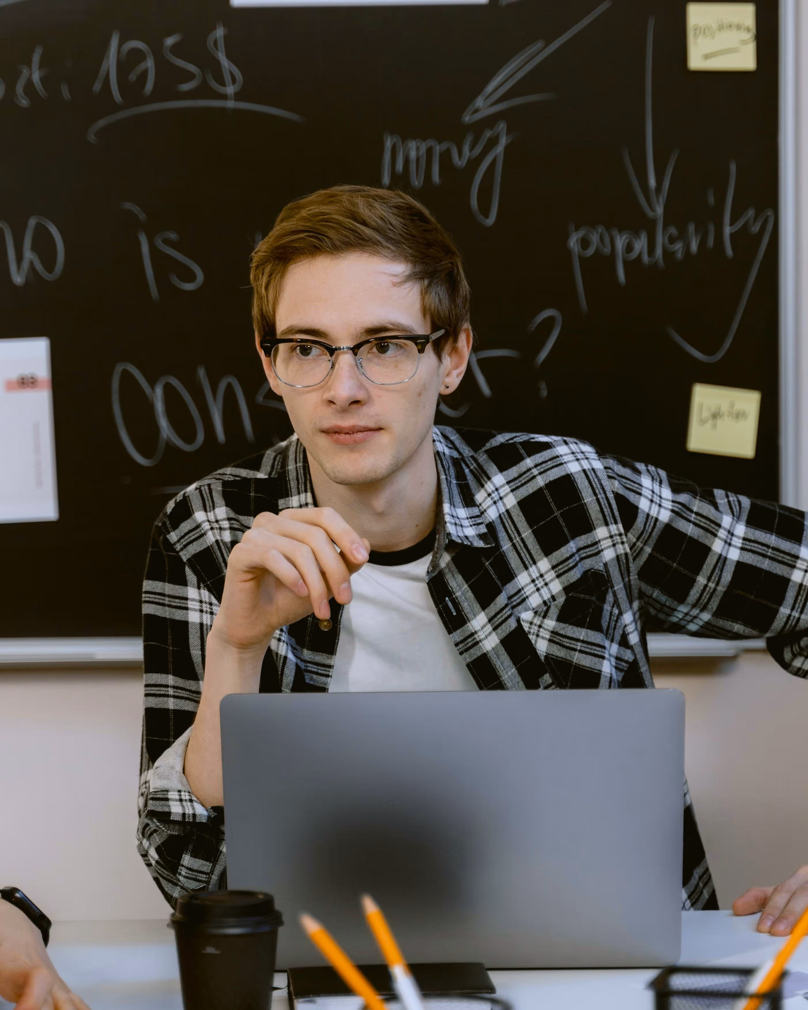 A young man in high school sitting in front of a computer at a table with lots of pencils and papers strewn about. He is holding a pen thoughtfully while listening to one of his teammates sitting on his right side.
