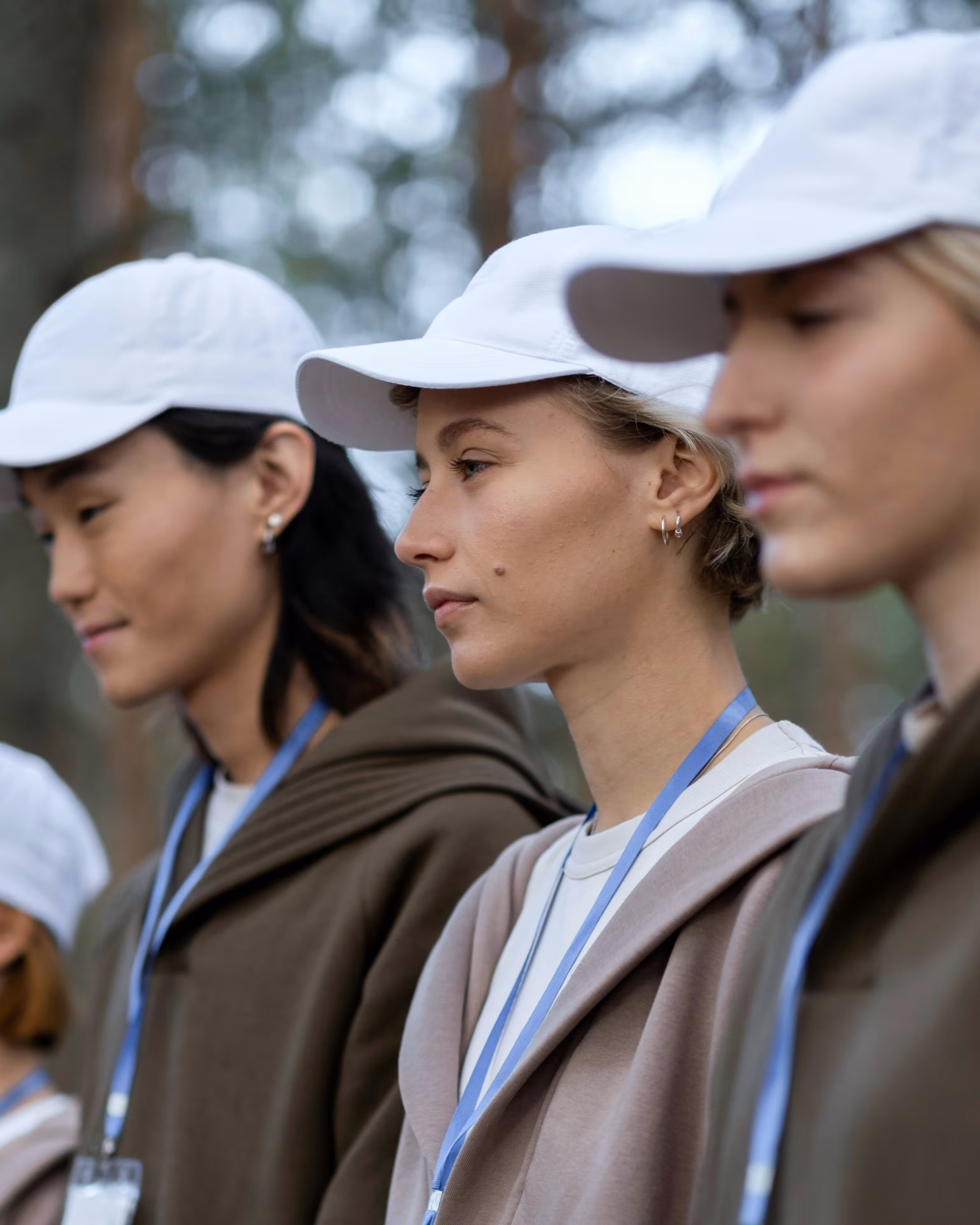A group of teenage girls wearing white hats, comfortable hoodies, and blue lanyards standing single-file and waiting for action. They are volunteering for an activity in a forested area. The main girl in focus stands in the center, with a determined stare facing forward.