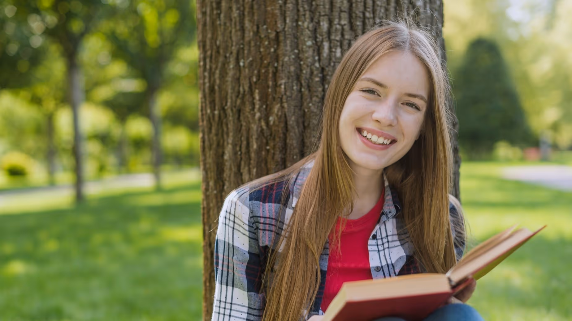 A young lady sitting in a field underneath the shade of a large tree. She is looking at the camera with a wide grin on her face and is holding a book open on her lap.