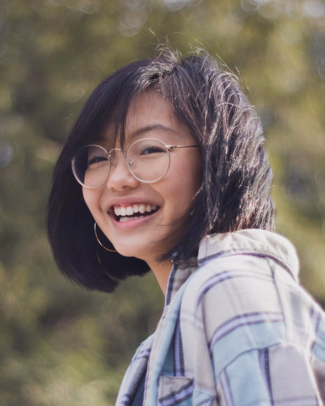 A young girl with rounded glasses turning to the side with a big smile on her face, looking at the camera.